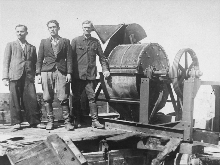 Jewish prisoners forced to work for a Sonderkommando 1005 unit pose next to a bone crushing machine in the Janowska concentration camp. Pictured from left to right are: unknown, David Manusevitz, and Moses Korn.